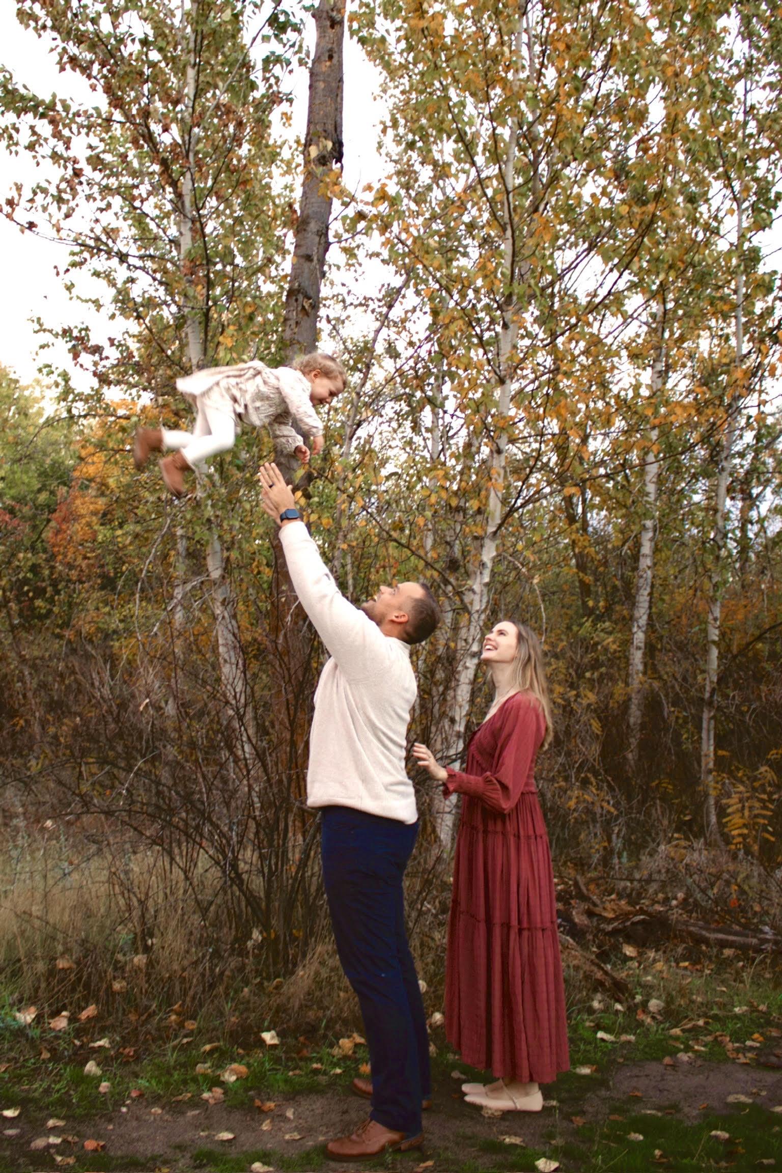 Lauren and her husband tossing their toddler into the air among golden autumn trees
